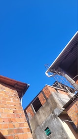 Two buildings with exposed brick and concrete walls appear under a clear, vivid blue sky. The roof of one building is partially covered with corrugated metal sheets, and there is a metal frame or antenna on the edge of the structure.