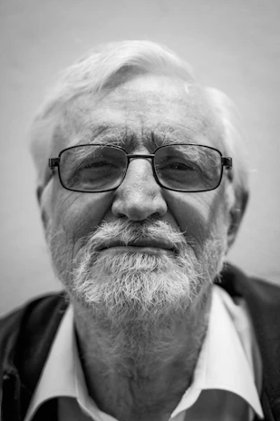 An elderly man with white hair and a beard, wearing glasses and a collared shirt under a dark jacket, is captured in a close-up black and white photo.