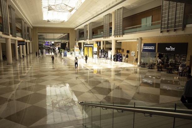A spacious, modern indoor hall with a high ceiling and large glass windows, featuring various people walking across a polished, checkered floor. The hall is lined with informational booths and small businesses, including a cafe area on the right. Bright lights and reflections illuminate the space, creating a sense of openness.