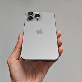 A hand is holding a modern smartphone with a sleek design. The back of the phone has a metallic finish and features three large camera lenses arranged in a triangular formation. There is a prominent logo in the center of the phone.