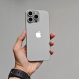 A hand is holding a modern smartphone with a sleek design. The phone has a gray finish with a prominent Apple logo on the back. It features three large camera lenses at the top left corner.