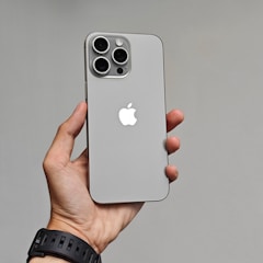 A hand is holding a modern smartphone with a sleek design. The phone has a gray finish with a prominent Apple logo on the back. It features three large camera lenses at the top left corner.