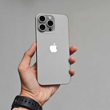 A hand is holding a modern smartphone with a sleek design. The phone has a gray finish with a prominent Apple logo on the back. It features three large camera lenses at the top left corner.