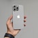 A hand is holding a modern smartphone with a sleek design. The phone has a gray finish with a prominent Apple logo on the back. It features three large camera lenses at the top left corner.