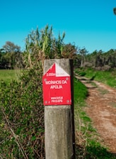 A wooden post with a red and white trail marker indicating a path named Moinhos da Apúlia is seen. The marker provides distance (2050 meters) and estimated walking time (54 minutes). The post is surrounded by greenery with tall grasses and trees in the background. A dirt pathway extends into the distance on the right side of the image under a clear blue sky.