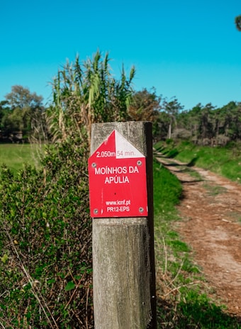 A wooden post with a red and white trail marker indicating a path named Moinhos da Apúlia is seen. The marker provides distance (2050 meters) and estimated walking time (54 minutes). The post is surrounded by greenery with tall grasses and trees in the background. A dirt pathway extends into the distance on the right side of the image under a clear blue sky.