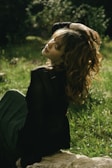 A person with curly hair sits outdoors on a stone, surrounded by green grass and lush foliage. The sunlight creates a warm glow on the person's hair and face. Their eyes are closed and a serene expression suggests tranquility.