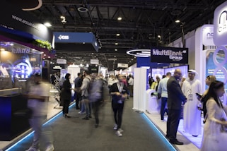 A bustling trade show or exposition with numerous booths displaying technology and financial services. People are interacting and walking around the exhibits, giving the scene a dynamic and busy atmosphere.