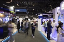 A bustling trade show or exposition with numerous booths displaying technology and financial services. People are interacting and walking around the exhibits, giving the scene a dynamic and busy atmosphere.
