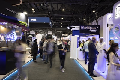 A bustling trade show or exposition with numerous booths displaying technology and financial services. People are interacting and walking around the exhibits, giving the scene a dynamic and busy atmosphere.
