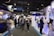 A bustling trade show or exposition with numerous booths displaying technology and financial services. People are interacting and walking around the exhibits, giving the scene a dynamic and busy atmosphere.
