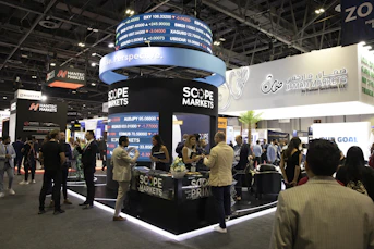 A bustling exhibition hall filled with people attending a financial conference or trade event. Prominent displays and booths from companies like Scope Markets, Daman Markets, and Hantec Markets are visible. Attendees are engaged in conversations, networking, and exploring the offerings and information provided by the exhibitors.