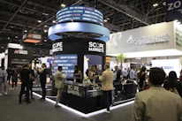 A bustling exhibition hall filled with people attending a financial conference or trade event. Prominent displays and booths from companies like Scope Markets, Daman Markets, and Hantec Markets are visible. Attendees are engaged in conversations, networking, and exploring the offerings and information provided by the exhibitors.