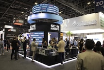 A bustling exhibition hall filled with people attending a financial conference or trade event. Prominent displays and booths from companies like Scope Markets, Daman Markets, and Hantec Markets are visible. Attendees are engaged in conversations, networking, and exploring the offerings and information provided by the exhibitors.