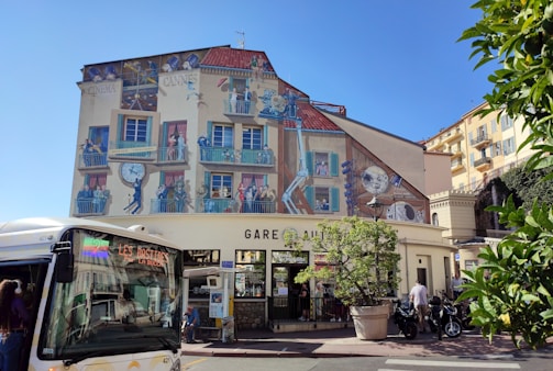 A vibrant building facade features a detailed mural depicting scenes related to cinema, with people engaged in various activities like filming and watching movies. The building is located in a sunny urban setting with a large clock as part of the mural. In front of the building, a bus is parked, and several scooters are nearby. There are trees and foliage adding a touch of greenery.