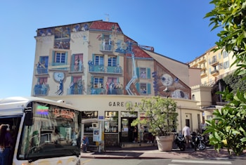 A vibrant building facade features a detailed mural depicting scenes related to cinema, with people engaged in various activities like filming and watching movies. The building is located in a sunny urban setting with a large clock as part of the mural. In front of the building, a bus is parked, and several scooters are nearby. There are trees and foliage adding a touch of greenery.