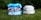 Two stacks of diapers are placed on grass, one stack features colorful, patterned reusable cloth diapers, and the other consists of plain white disposable diapers. The background includes green foliage.