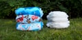 Two stacks of diapers are placed on grass, one stack features colorful, patterned reusable cloth diapers, and the other consists of plain white disposable diapers. The background includes green foliage.