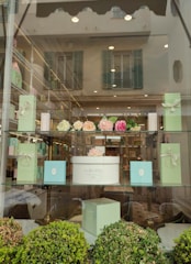 The image features a window display of a boutique or patisserie with elegant boxes in pastel green and a white box labeled 'Ladurée Paris'. Above the box are artificial flower arrangements in shades of pink and cream. The display is set in a well-lit store with reflective glass showing an exterior building with shutters.