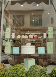 The image features a window display of a boutique or patisserie with elegant boxes in pastel green and a white box labeled 'Ladur&eacute;e Paris'. Above the box are artificial flower arrangements in shades of pink and cream. The display is set in a well-lit store with reflective glass showing an exterior building with shutters.