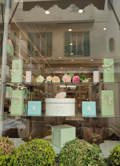 The image features a window display of a boutique or patisserie with elegant boxes in pastel green and a white box labeled 'Ladur&eacute;e Paris'. Above the box are artificial flower arrangements in shades of pink and cream. The display is set in a well-lit store with reflective glass showing an exterior building with shutters.