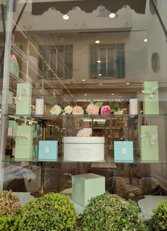 The image features a window display of a boutique or patisserie with elegant boxes in pastel green and a white box labeled 'Ladur&eacute;e Paris'. Above the box are artificial flower arrangements in shades of pink and cream. The display is set in a well-lit store with reflective glass showing an exterior building with shutters.