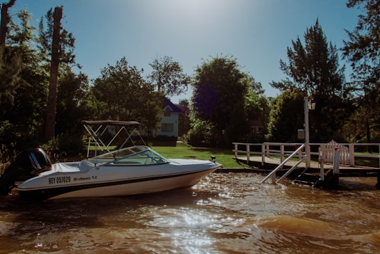 A white motorboat is docked by a wooden pier on a calm, brown river. The background features lush green trees and a white house with blue accents partially obscured by foliage. The scene is illuminated by bright sunlight.