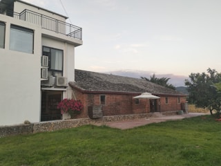 A rustic brick building with a sloped roof next to a modern white structure featuring large windows and several air conditioning units. A patio umbrella stands in the courtyard, adjacent to neatly maintained green grass and shrubbery. The sky is mostly clear, and distant mountains are visible in the background.