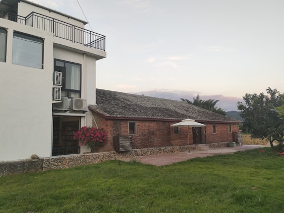A rustic brick building with a sloped roof next to a modern white structure featuring large windows and several air conditioning units. A patio umbrella stands in the courtyard, adjacent to neatly maintained green grass and shrubbery. The sky is mostly clear, and distant mountains are visible in the background.