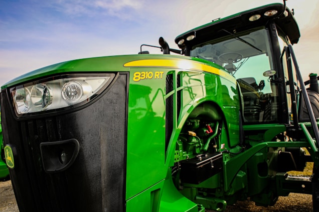 A large green agricultural tractor with the model number 8310 RT prominently displayed on its side. The vehicle features a robust build, with a visible engine compartment and modern headlights. The driver’s cabin is visible with clear windows, and the tractor is parked on a gravel surface under a partly cloudy sky.