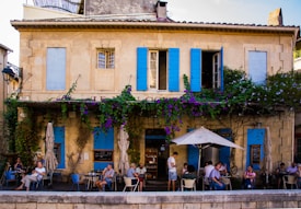 A quaint caf&eacute; with blue shutters and a warm yellow facade. Vines with purple flowers climb the walls, creating a cozy atmosphere. Several people are seated at outdoor tables under umbrellas, engaged in conversation. The ambiance is relaxed and inviting, suggesting a typical street scene in a small European town.