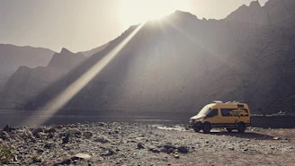 A sunlit campervan parked by a serene lakeside at dawn, with soft mist rising from the water.