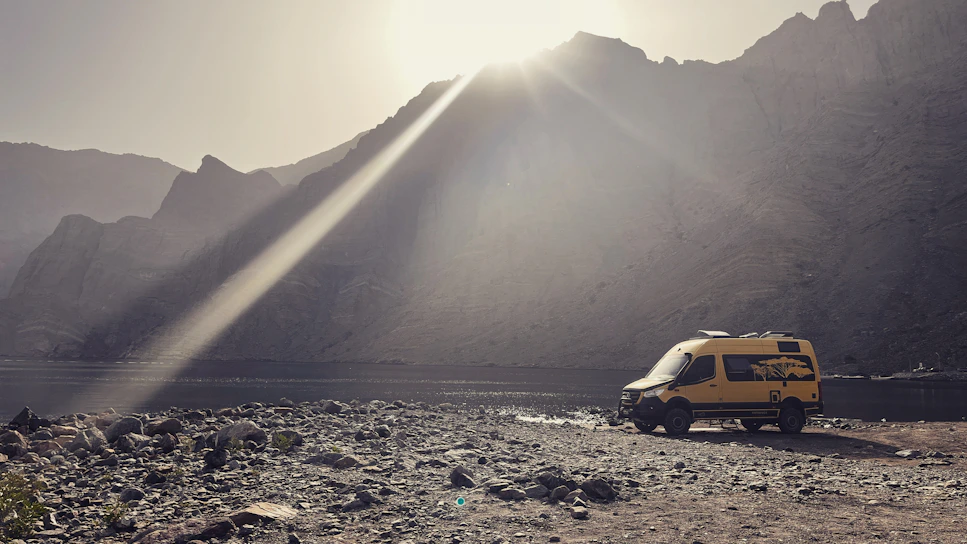 A sunlit campervan parked by a serene lakeside at dawn, with soft mist rising from the water.