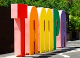 Large, colorful three-dimensional letters spell out the word 'Tampa' in an outdoor setting, with greenery in the background and bright sunlight casting shadows.