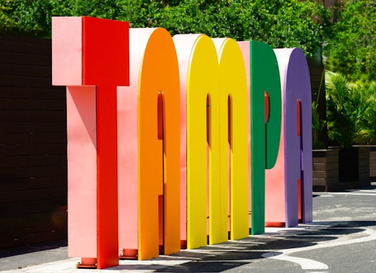 Large, colorful three-dimensional letters spell out the word 'Tampa' in an outdoor setting, with greenery in the background and bright sunlight casting shadows.