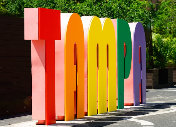 Large, colorful three-dimensional letters spell out the word 'Tampa' in an outdoor setting, with greenery in the background and bright sunlight casting shadows.