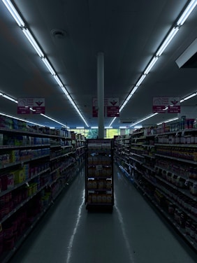 A dimly lit aisle in a grocery store with shelves on both sides filled with various products. The aisle is empty and illuminated by fluorescent lights. The signs above the aisles provide information about the sections, indicating areas for farm fresh foods, along with beverages and snacks.