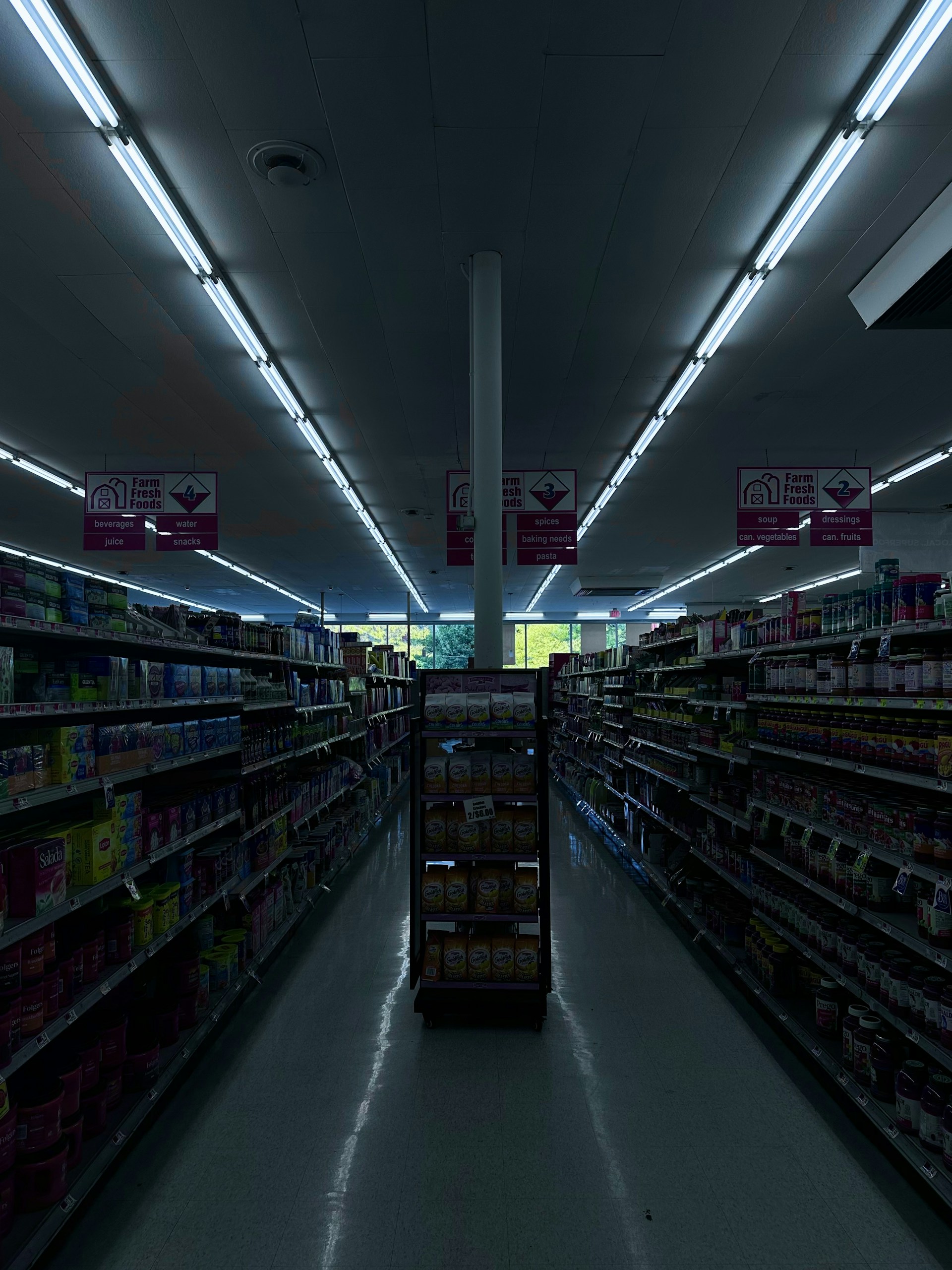 A dimly lit aisle in a grocery store with shelves on both sides filled with various products. The aisle is empty and illuminated by fluorescent lights. The signs above the aisles provide information about the sections, indicating areas for farm fresh foods, along with beverages and snacks.