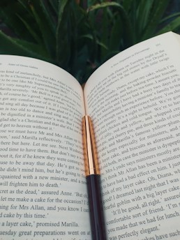 An open book with a black and gold fountain pen placed on it. The background is filled with green foliage, suggesting an outdoor setting.