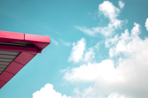 A part of a building's roof with red and gray metal panels extends into a bright blue sky filled with fluffy white clouds. The architecture is modern and minimalistic, with clean lines contrasting against the natural backdrop.