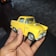 A hand is holding a small yellow toy car, which appears to be a model of an old-fashioned vehicle. The car has a distinctive design with a visible chrome front bumper, detailed front grille, and hood ornament. The toy car is well-detailed, with visible windows, tires, and door lines. The background consists of a dark, blurred surface, possibly a floor or table.