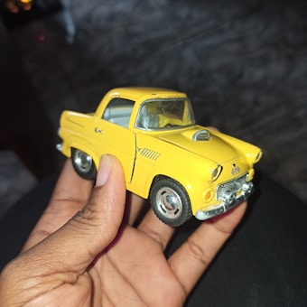 A hand is holding a small yellow toy car, which appears to be a model of an old-fashioned vehicle. The car has a distinctive design with a visible chrome front bumper, detailed front grille, and hood ornament. The toy car is well-detailed, with visible windows, tires, and door lines. The background consists of a dark, blurred surface, possibly a floor or table.