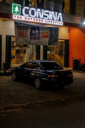 A dark-colored car is parked in front of an outdoor lifestyle store named Consina. The store has a lit sign with a green symbol and white letters. A large banner advertising a payday sale with up to 50% off is hanging above the car. The storefront has large glass windows with a clear view into the interior, which appears to contain various sporting goods and outdoor equipment. A scooter is parked to the right side of the car near the store entrance.