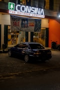 A dark-colored car is parked in front of an outdoor lifestyle store named Consina. The store has a lit sign with a green symbol and white letters. A large banner advertising a payday sale with up to 50% off is hanging above the car. The storefront has large glass windows with a clear view into the interior, which appears to contain various sporting goods and outdoor equipment. A scooter is parked to the right side of the car near the store entrance.