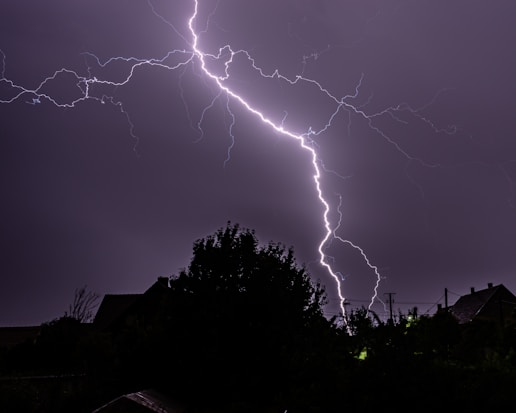 A dramatic lightning bolt illuminates the night sky, branching across the dark clouds with a backdrop of silhouetted trees and rooftops. The scene captures the raw power and beauty of a storm.