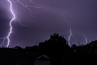 Multiple bright lightning strikes illuminate a dark, cloudy night sky, silhouetting the landscape below, including trees and rooftops.