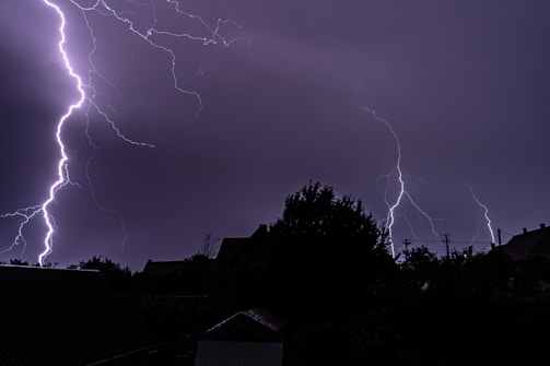 Multiple bright lightning strikes illuminate a dark, cloudy night sky, silhouetting the landscape below, including trees and rooftops.