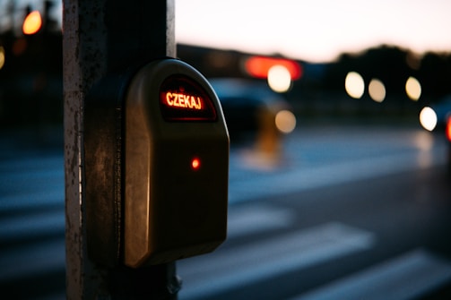 A pedestrian crossing button at dusk, with the word 'CZEKAJ' illuminated in red. The background is blurred, showing streaks of lights from cars on the road.