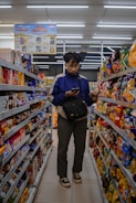 A person stands in the aisle of a store, browsing items on the shelves while looking at their phone. The shelves are filled with various snacks and packaged goods. Overhead fluorescent lights illuminate the space, and there is signage advertising discounts.