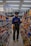 A person stands in the aisle of a store, browsing items on the shelves while looking at their phone. The shelves are filled with various snacks and packaged goods. Overhead fluorescent lights illuminate the space, and there is signage advertising discounts.